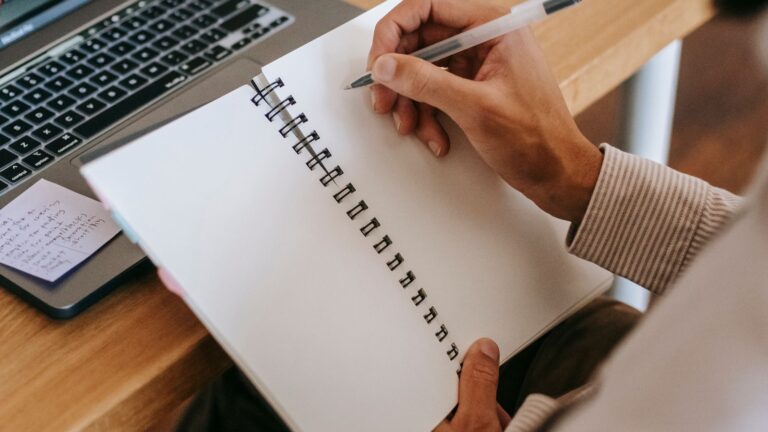 Man writing in a notebook beside a laptop and a cup of coffee in a home office while planning his work
